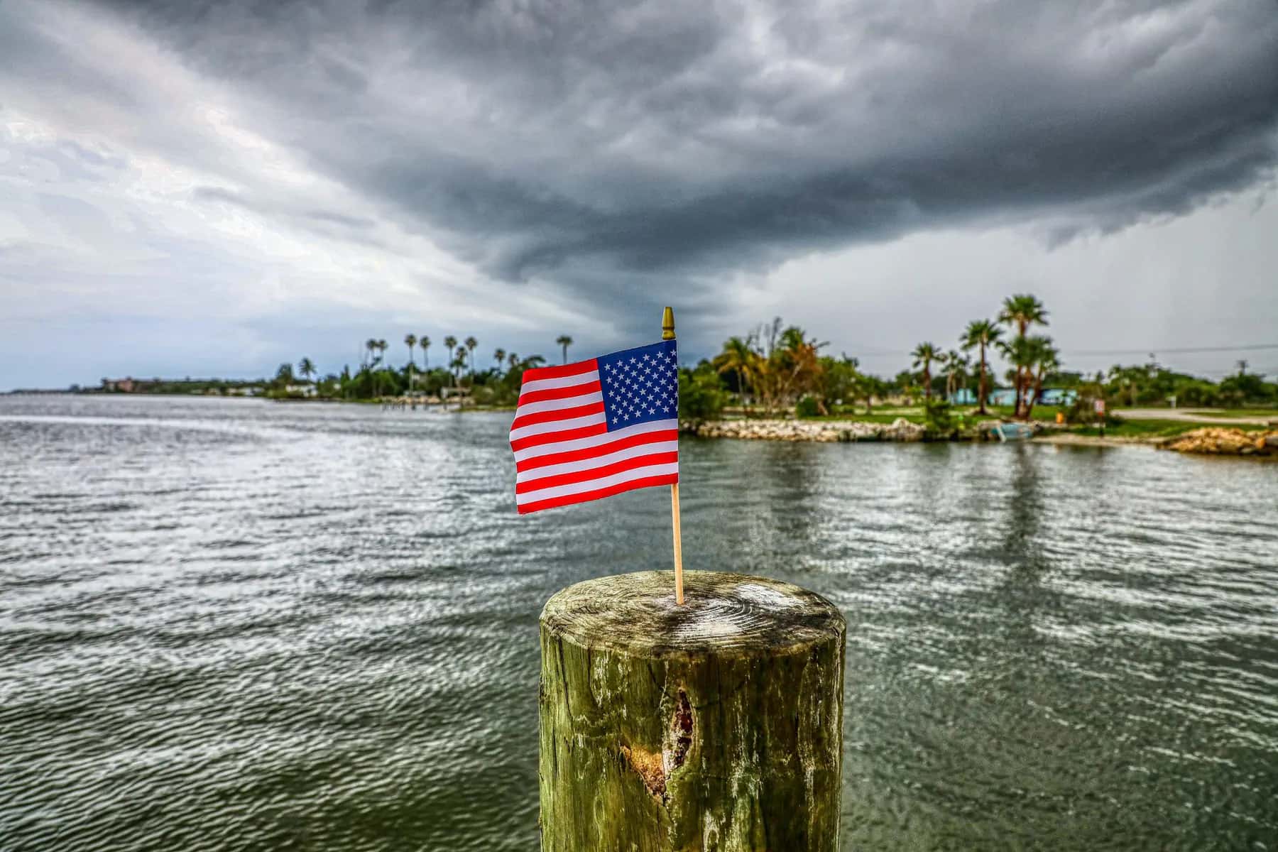 Dark storm clouds over a palm-lined shoreline with an American flag on a piling in the foreground