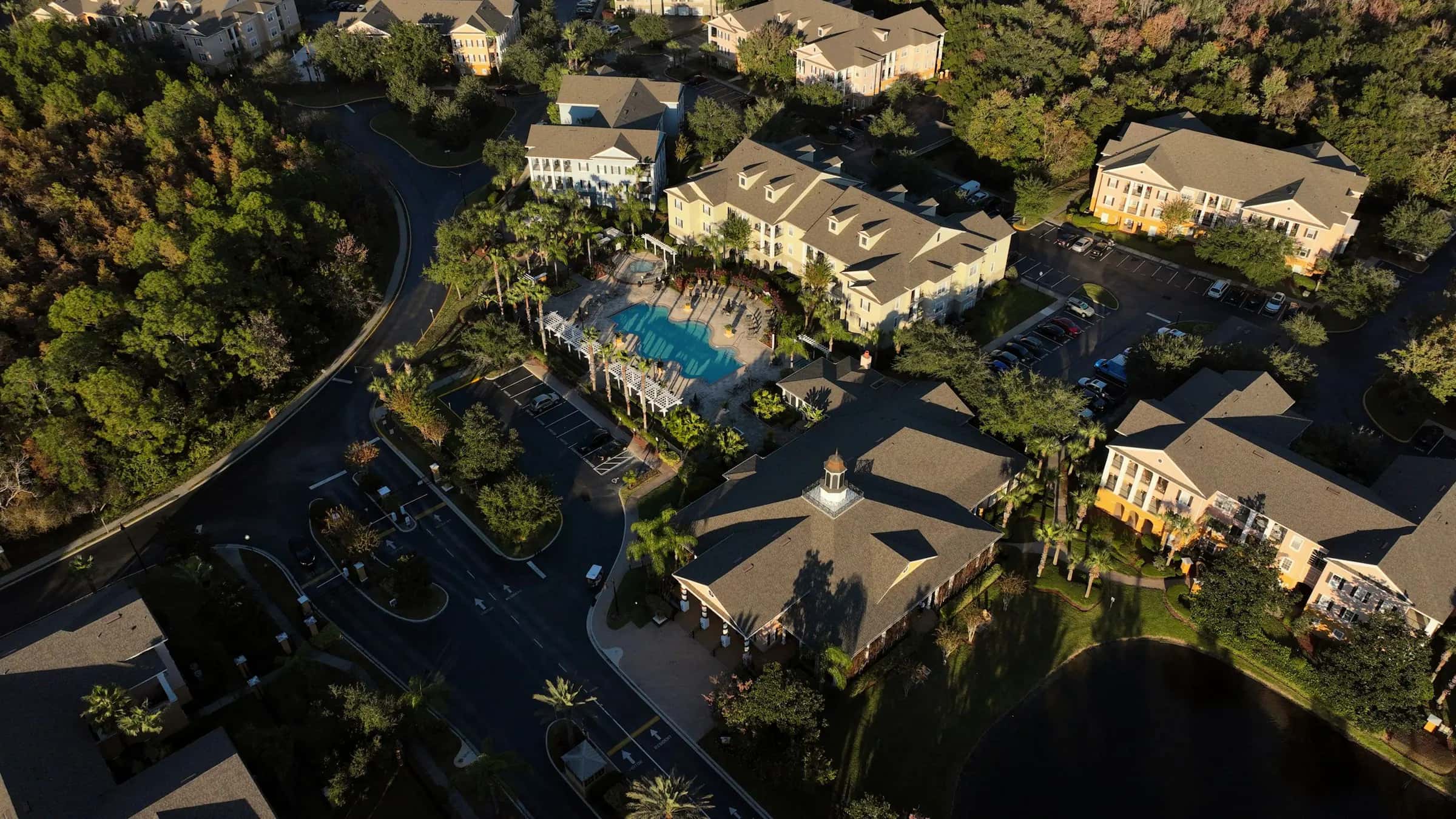 Aerial view at golden hour of a multi-building apartment community with a pool, palm trees, parking lots and a lake
