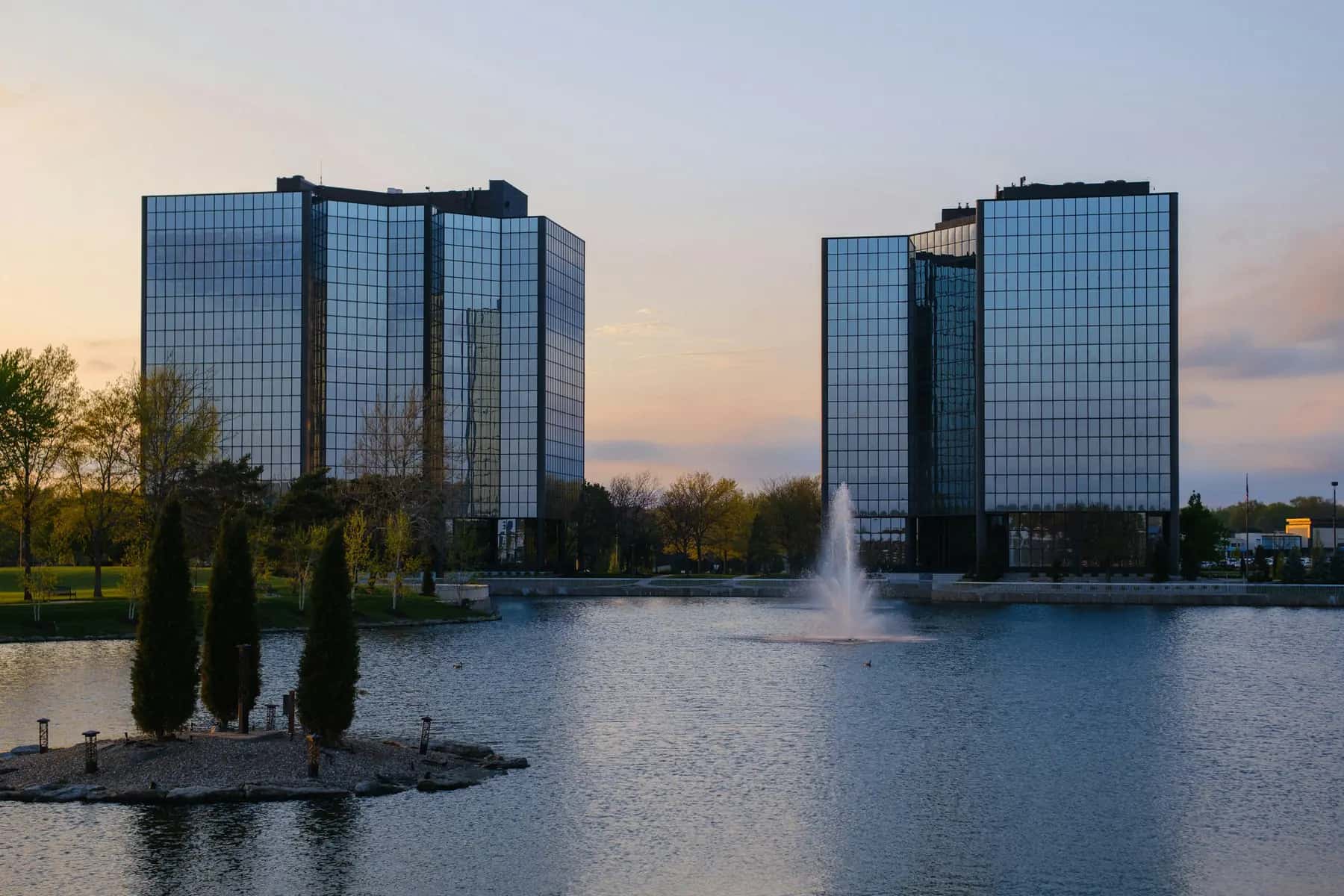 Two glass office towers reflected in a lake with a fountain at sunset