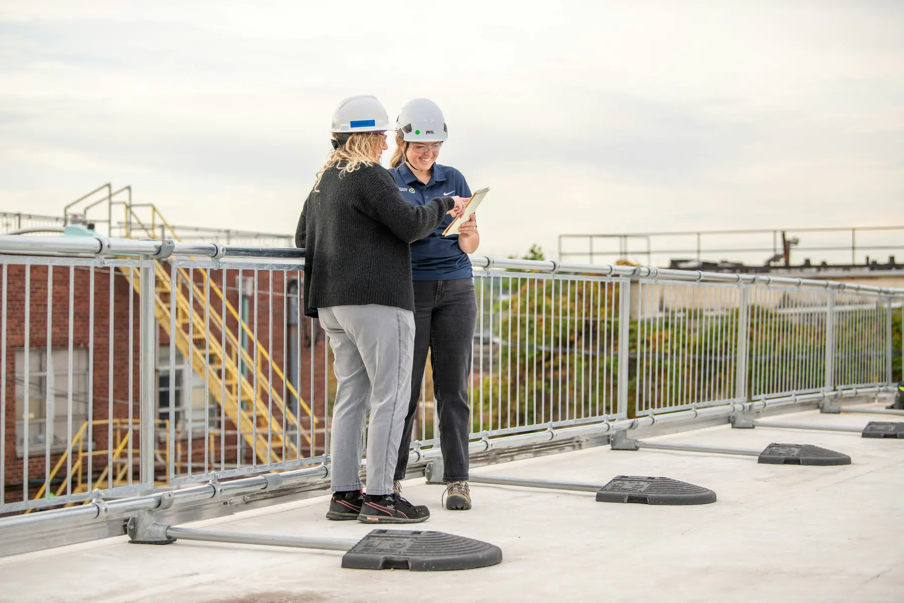 Two professionals in hard hats reviewing a tablet on a commercial rooftop with safety railings