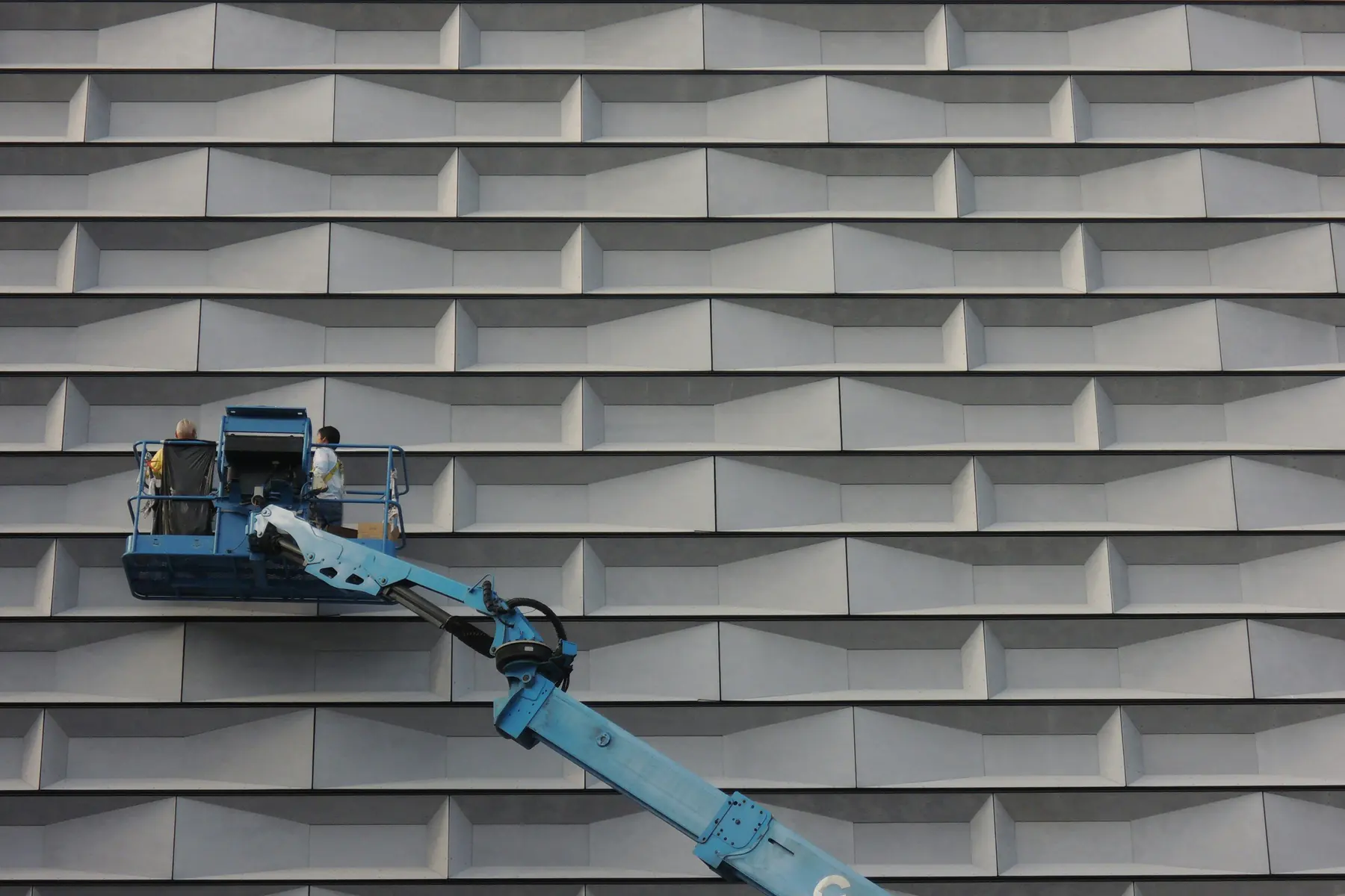 Two workers on a boom lift working on the gray paneled exterior wall of a large building