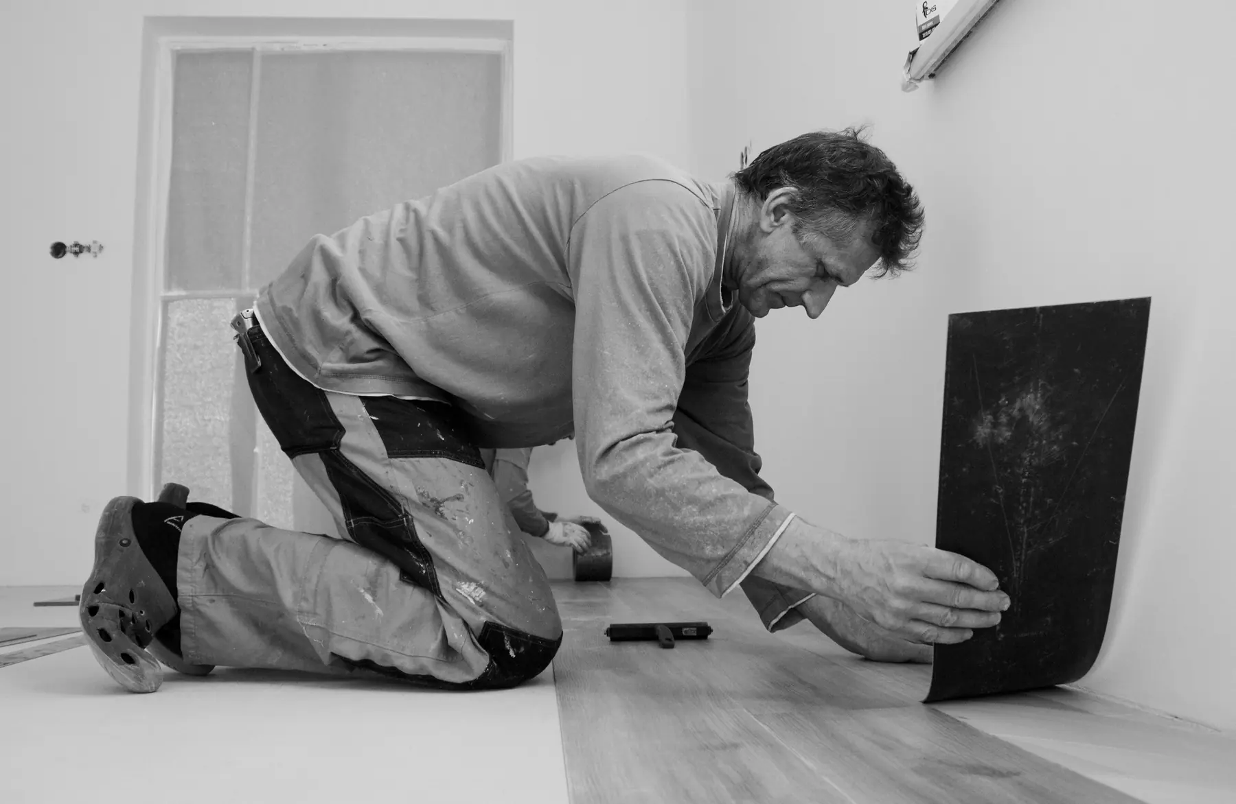 Installer kneeling to fit a vinyl plank against the wall of an empty room (black-and-white)