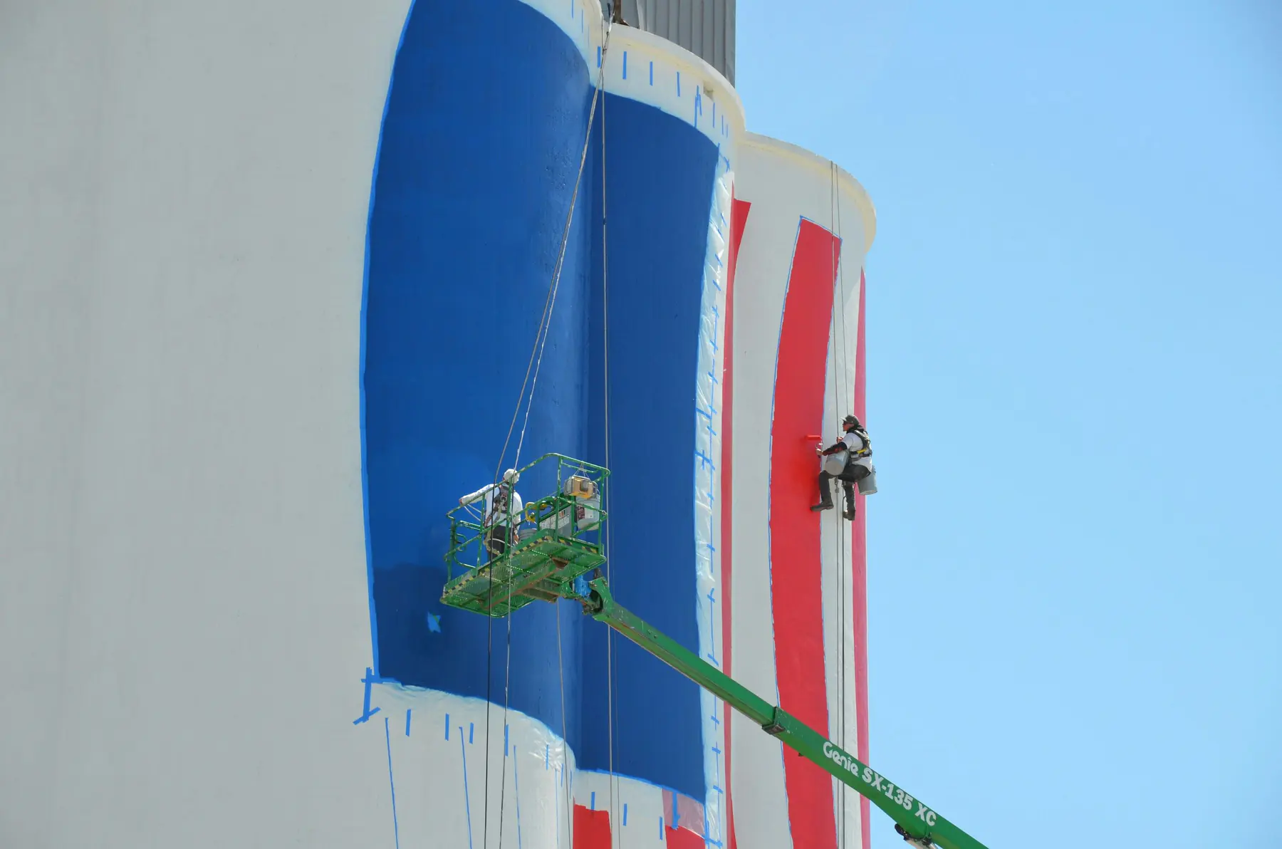 Painters on a boom lift and rope rigging painting a large mural on an industrial silo