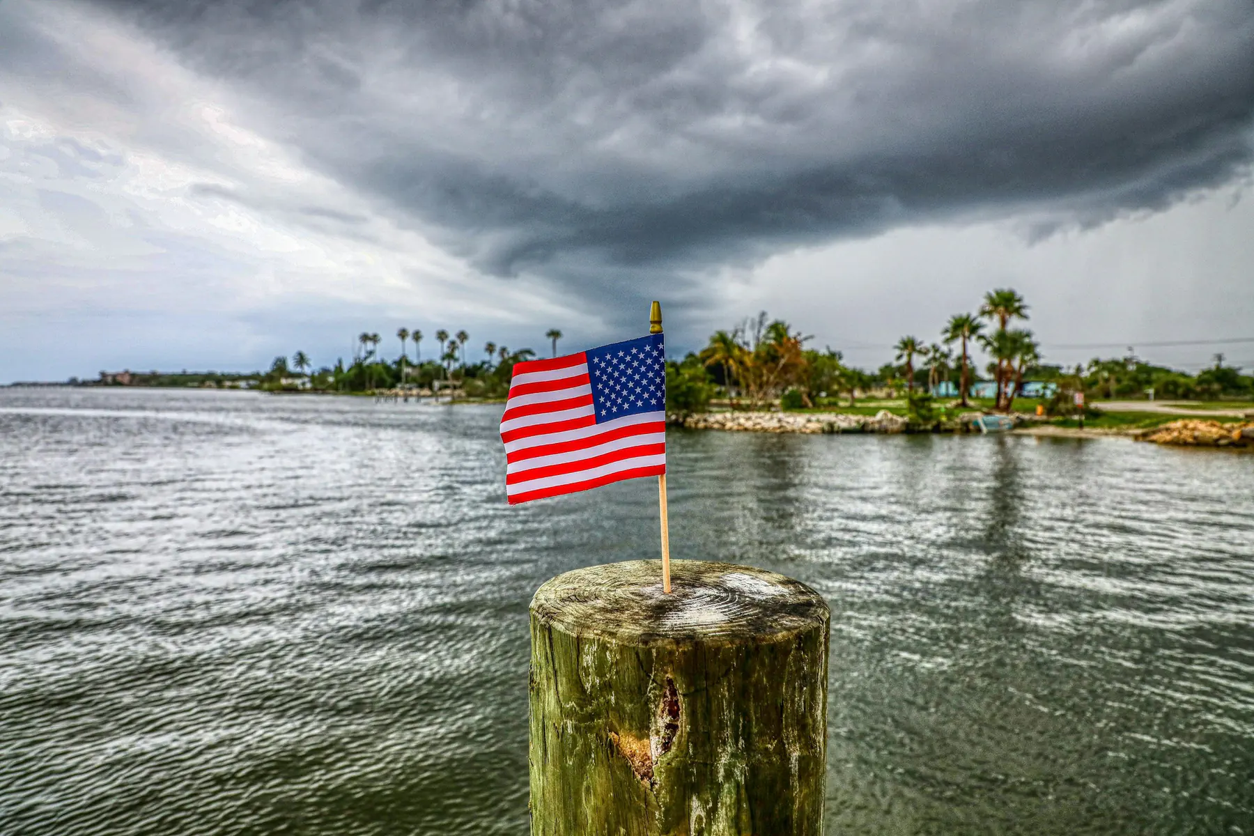 Dark storm clouds over a palm-lined shoreline with an American flag on a piling in the foreground