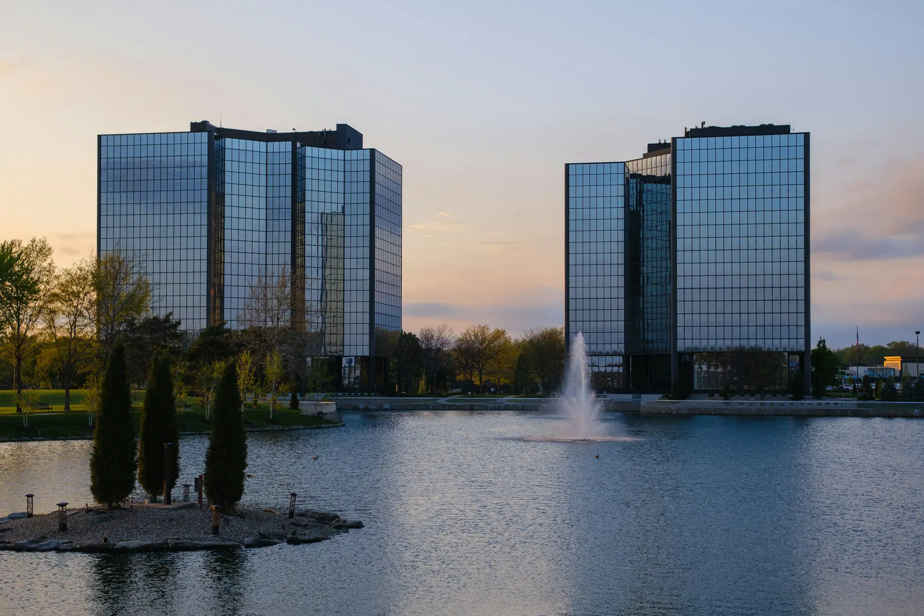 Two glass office towers reflected in a lake with a fountain at sunset