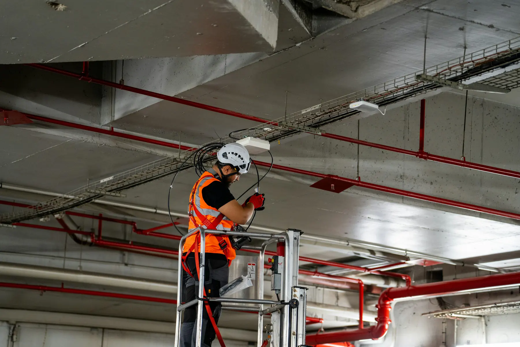Technician in a hard hat and hi-vis vest working from a lift on ceiling cabling in a parking garage