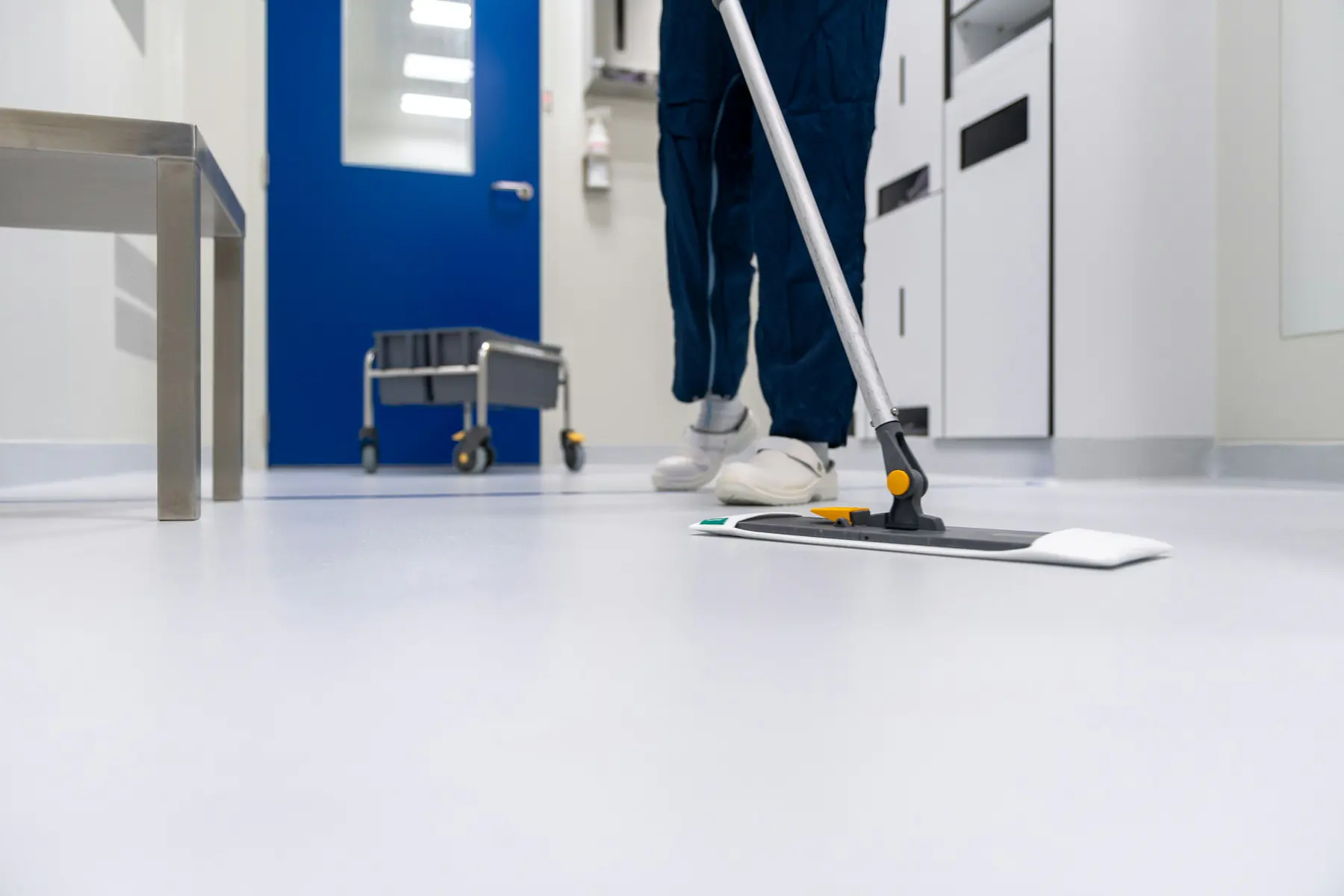 Worker in blue coveralls mopping a white floor in a clinical hallway