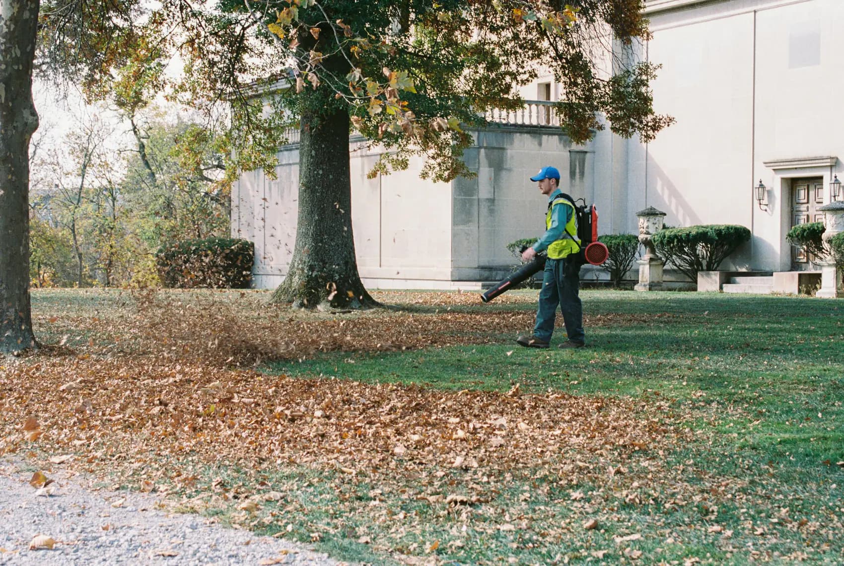 Groundskeeper in a hi-vis vest using a backpack leaf blower on a lawn beside a large building